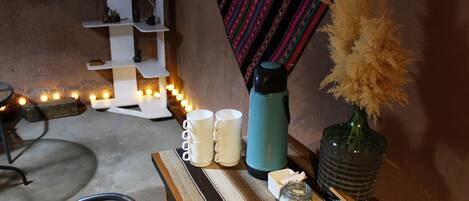 Un rincón acogedor con un sillón de mimbre, una mesita con una botella de agua azul, tazas blancas, un tarro de cristal y un jarrón decorativo con plantas secas.