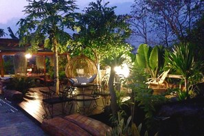 Un patio al aire libre con arreglos de asientos, incluyendo una silla colgante y un banco acolchado, rodeado de abundante follaje y una pérgola de madera.