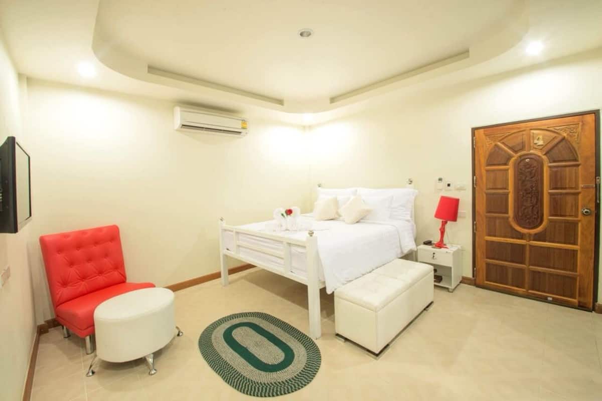 Un dormitorio con una cama blanca, una silla roja, un banco blanco, una puerta de madera y un equipo de aire acondicionado.