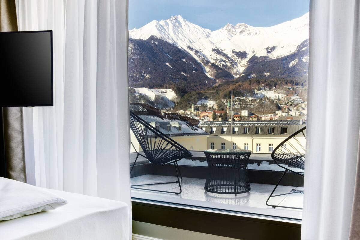 Een moderne hotelkamer met een groot raam met uitzicht op besneeuwde bergen en een stad eronder.