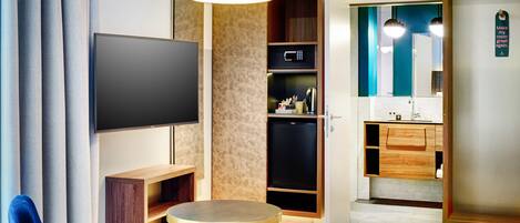 A modern hotel room with a flat-screen TV, a round table, a blue chair, a small ottoman, a microwave, a mini-fridge, a wooden cabinet, a vanity with a mirror, and a door with a sign.