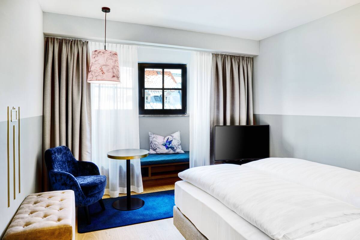 Een hotelkamer met een bed, een televisie, een blauwe stoel, een klein tafeltje en een raam met gordijnen.