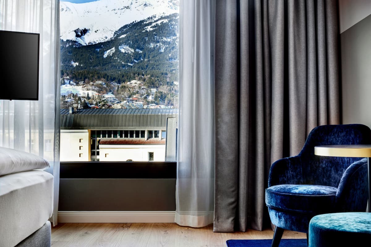 Een moderne hotelkamer met een groot raam dat uitzicht biedt op besneeuwde bergtoppen en een stad daaronder.