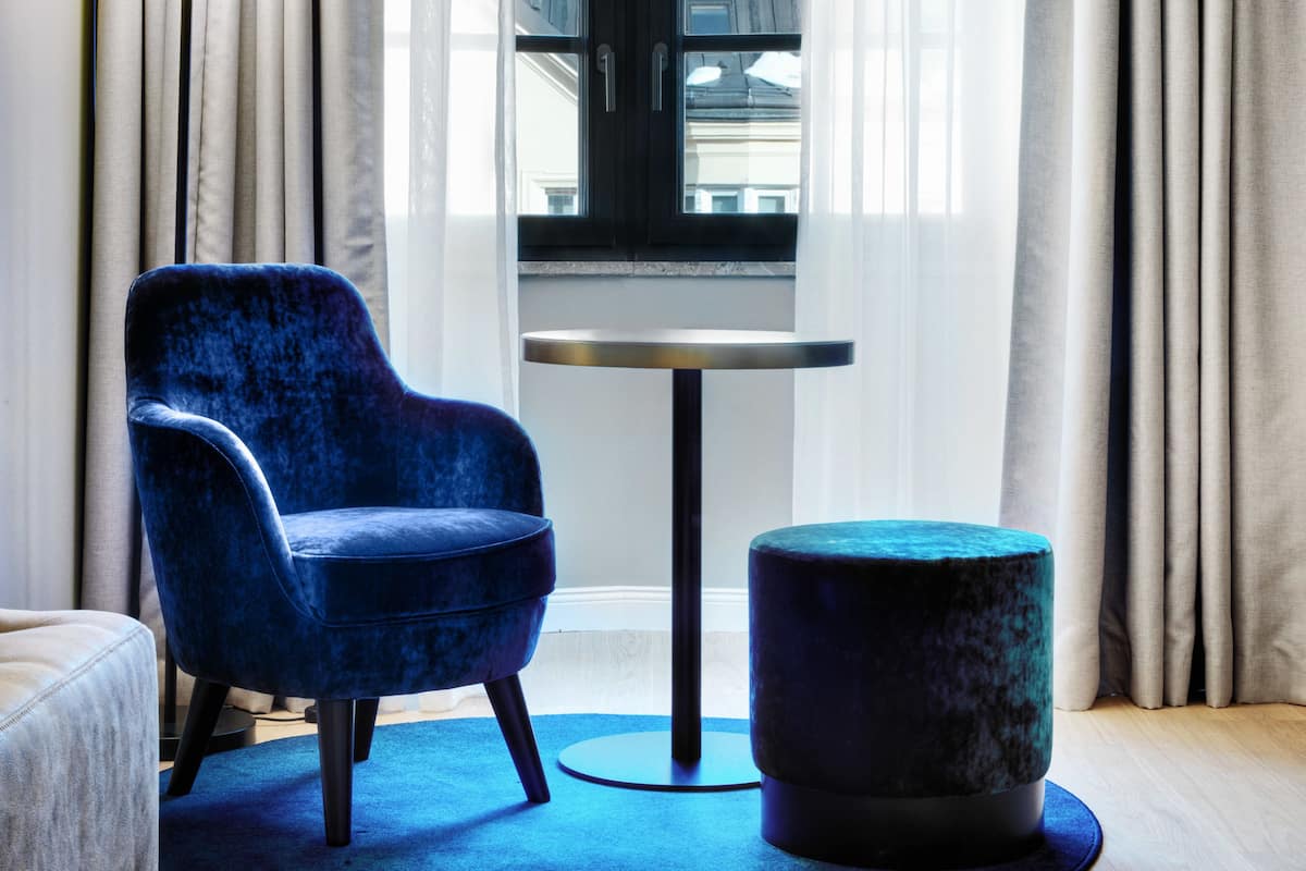 Een kamer met een blauwe fluweelstoel, een rond poefje, een klein rond tafeltje en een hanglamp met bloemenprint.
