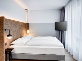A modern hotel room with a large bed, a flat-screen TV, a wall-mounted lamp, and a small table with a remote control.