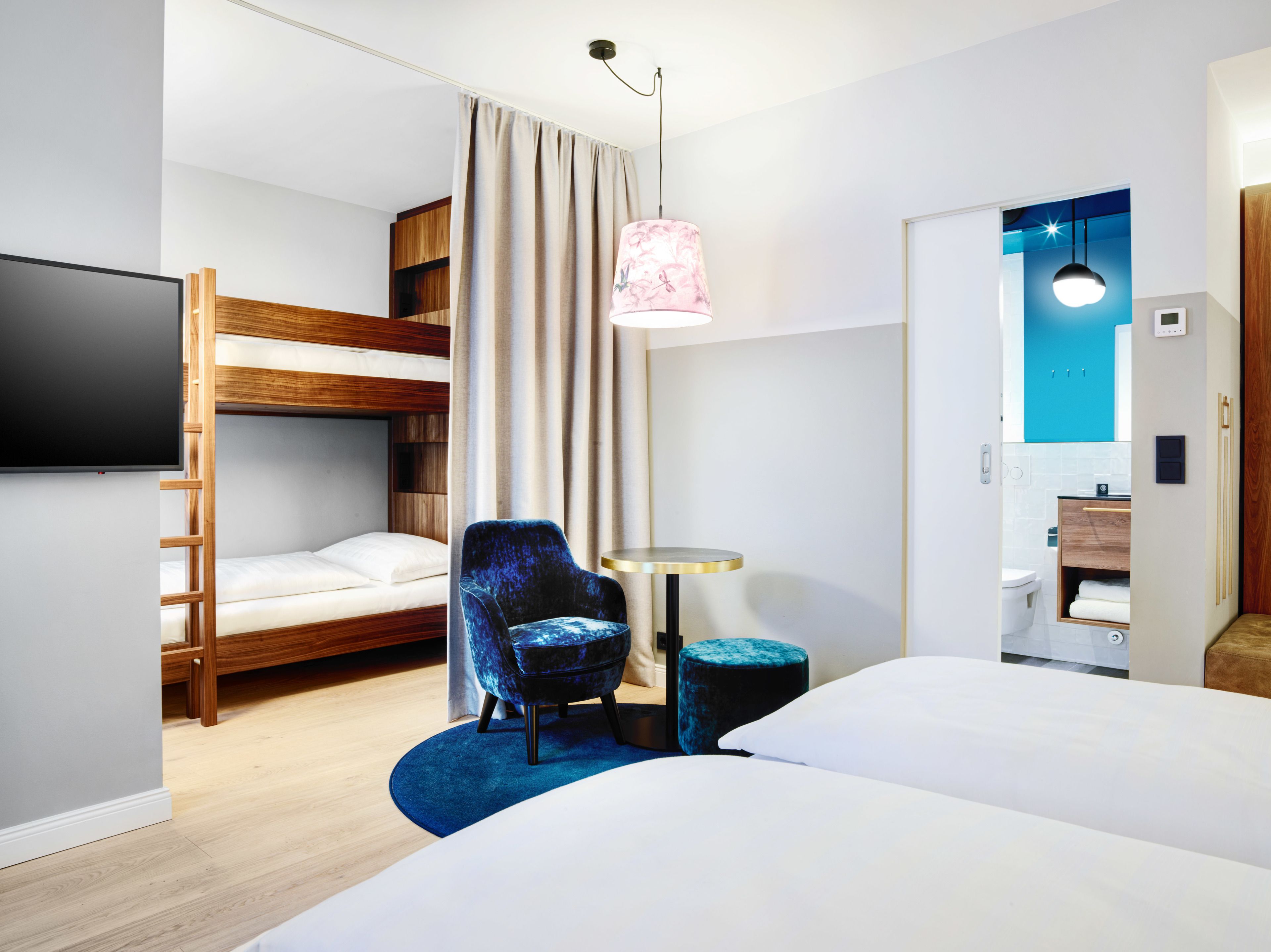 Une chambre d’hôtel moderne équipée d’un lit superposé, d’un lit simple, d’une table ronde, d’un fauteuil bleu, d’un petit pouf et d’un téléviseur fixé au mur.