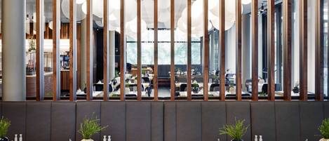 Un restaurante moderno con vistas a través de listones de madera, con mesas con manteles blancos, sillas negras y plantas en macetas.