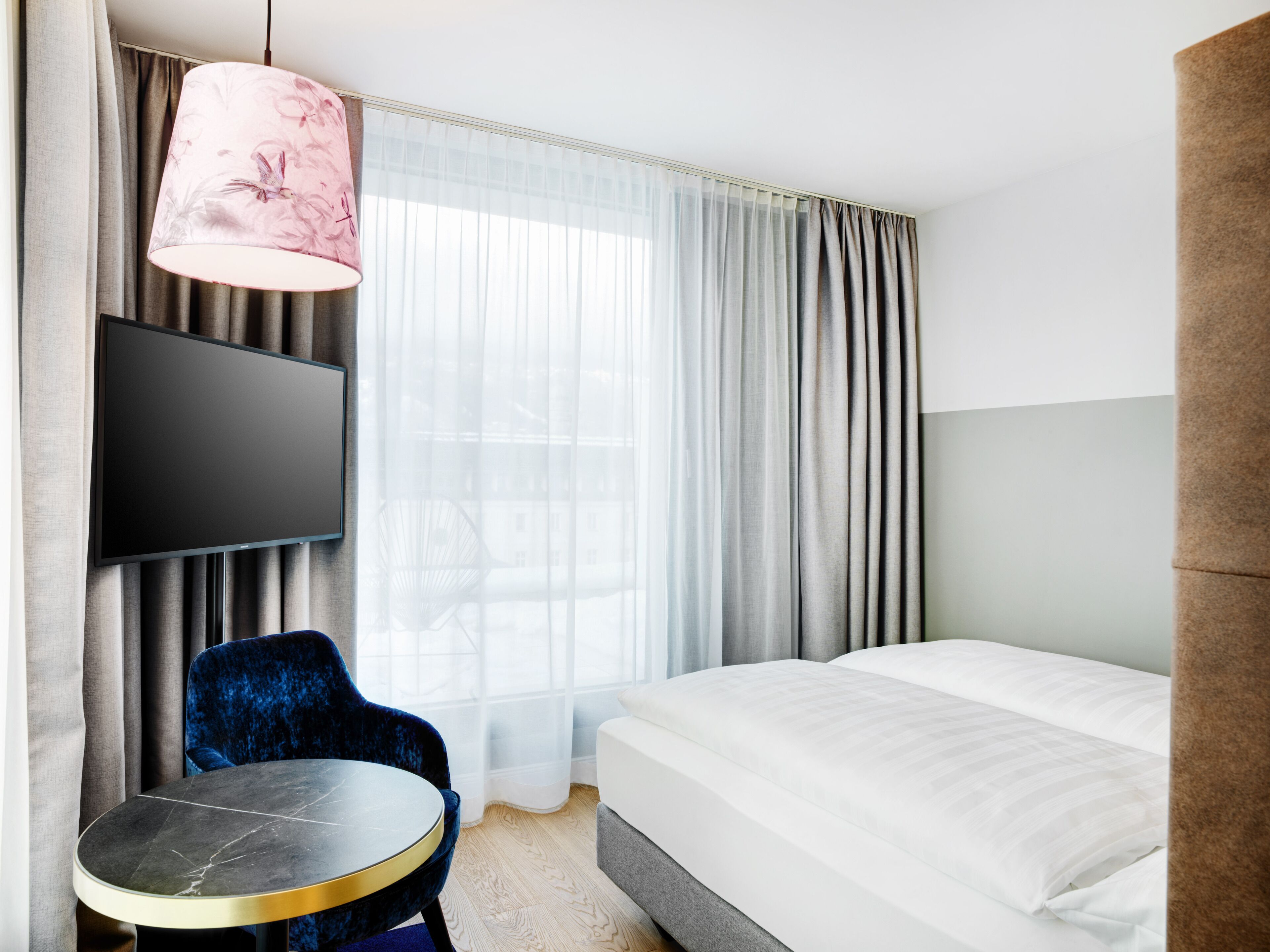 A hotel room with a bed, a TV mounted on the wall, a round table, a blue chair, and a pink lamp.