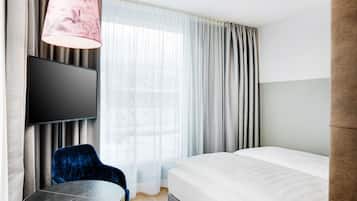 A hotel room with a bed, a TV mounted on the wall, a round table, a blue chair, and a pink lamp.