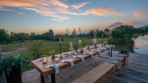 A wooden deck with a long table set for a meal, surrounded by candles and greenery, overlooking a body of water and a grassy field. - Gulls Way (The Crags)