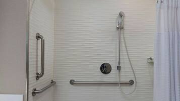 Ducha a nivel con puerta de vidrio, barras de apoyo, cabezal de ducha manual y cortina blanca.