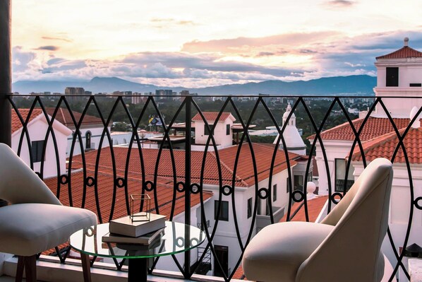 Un balcón con vistas a un paisaje urbano, que incluye edificios con tejados rojos y una torre blanca. Hay dos sillas blancas y una pequeña mesa redonda con un libro y un objeto de vidrio encima.