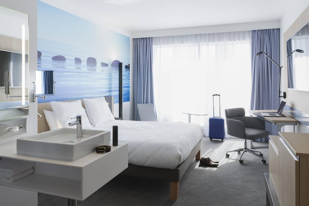 Hotellihuone, jossa on sänky, työpöytä kannettavalla tietokoneella, tuoli, matkalaukku, kylpyhuone, jossa on pesuallas ja peili, sekä suuri ikkuna, jossa on verhot.