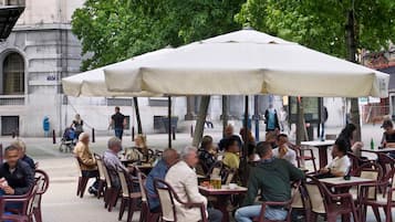 Un café al aire libre con mesas y sillas bajo un gran paraguas, rodeado de árboles y edificios.