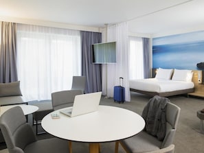 A hotel room with a bed, a TV, a desk with a laptop, a chair, and a suitcase. - Novotel Charleroi Centre (Charleroi)
