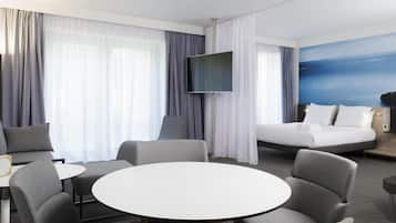 Quarto de hotel com uma cama, televisão, mesa redonda e duas poltronas.