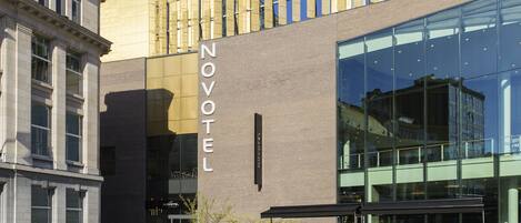 Un hotel moderno con un llamativo letrero de 'NOVOTEL', grandes ventanales y un área de estar al aire libre.
