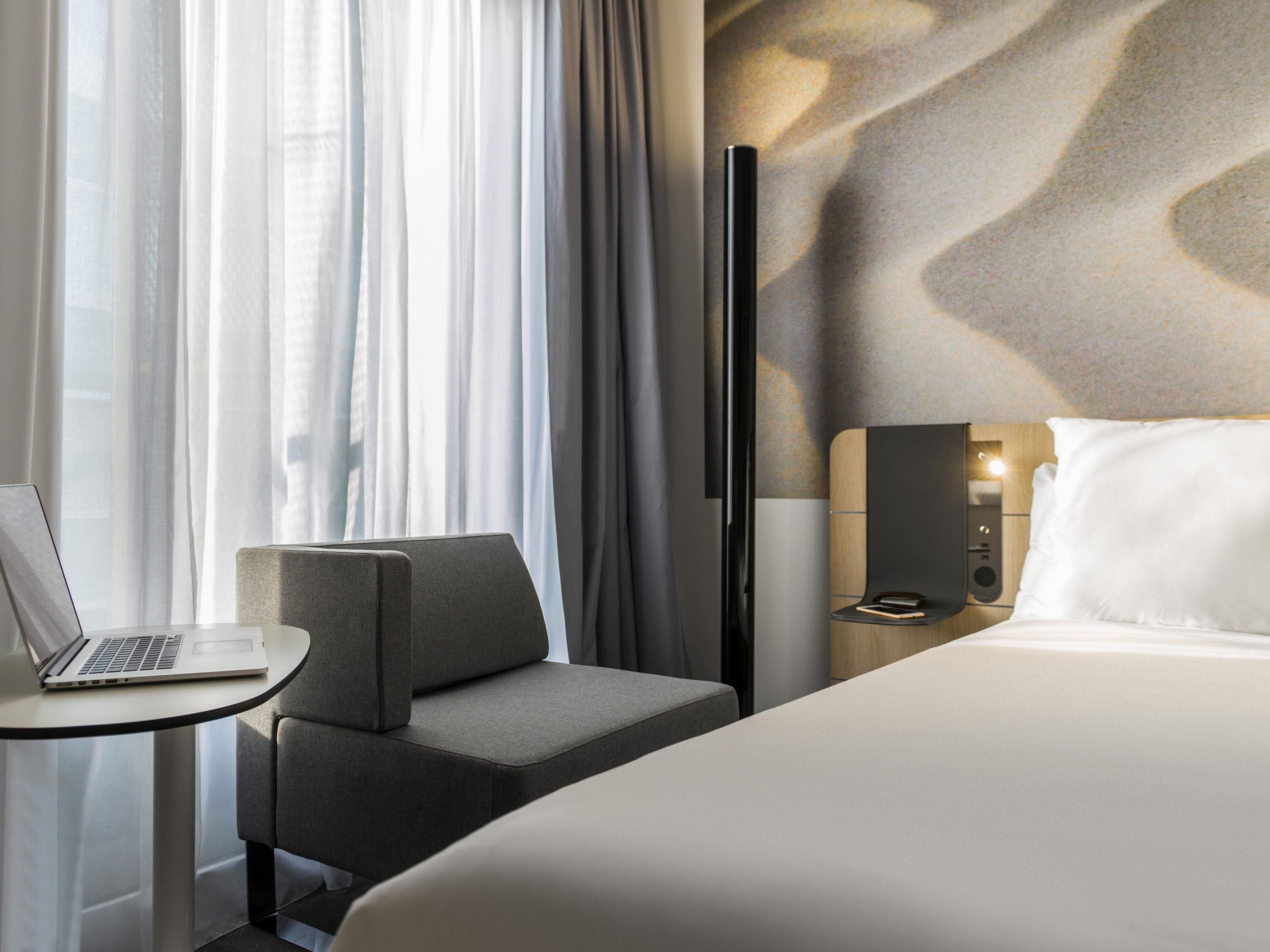 Habitación de hotel con cama, escritorio con computadora portátil, silla, mesita de noche y televisor de pantalla plana montado en la pared.
