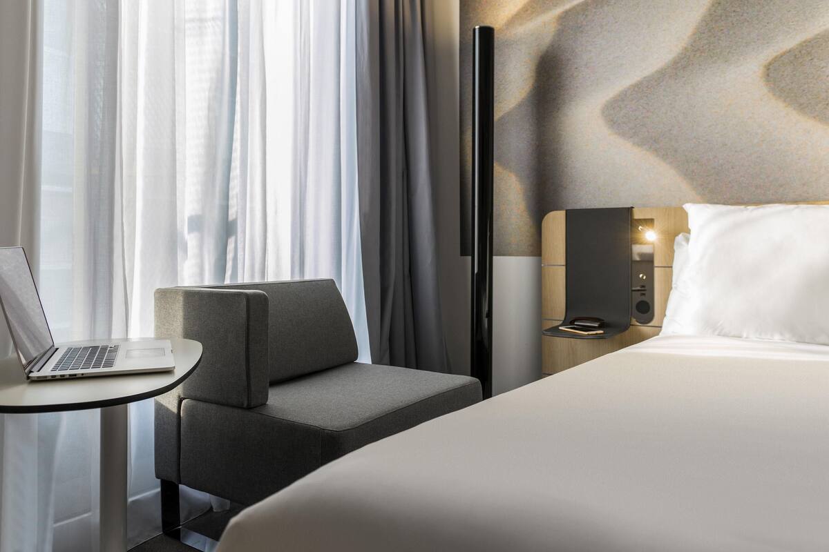 Hotellihuone, jossa on sänky, työpöytä kannettavalla tietokoneella, tuoli, yöpöytä ja seinälle asennettu taulutelevisio.