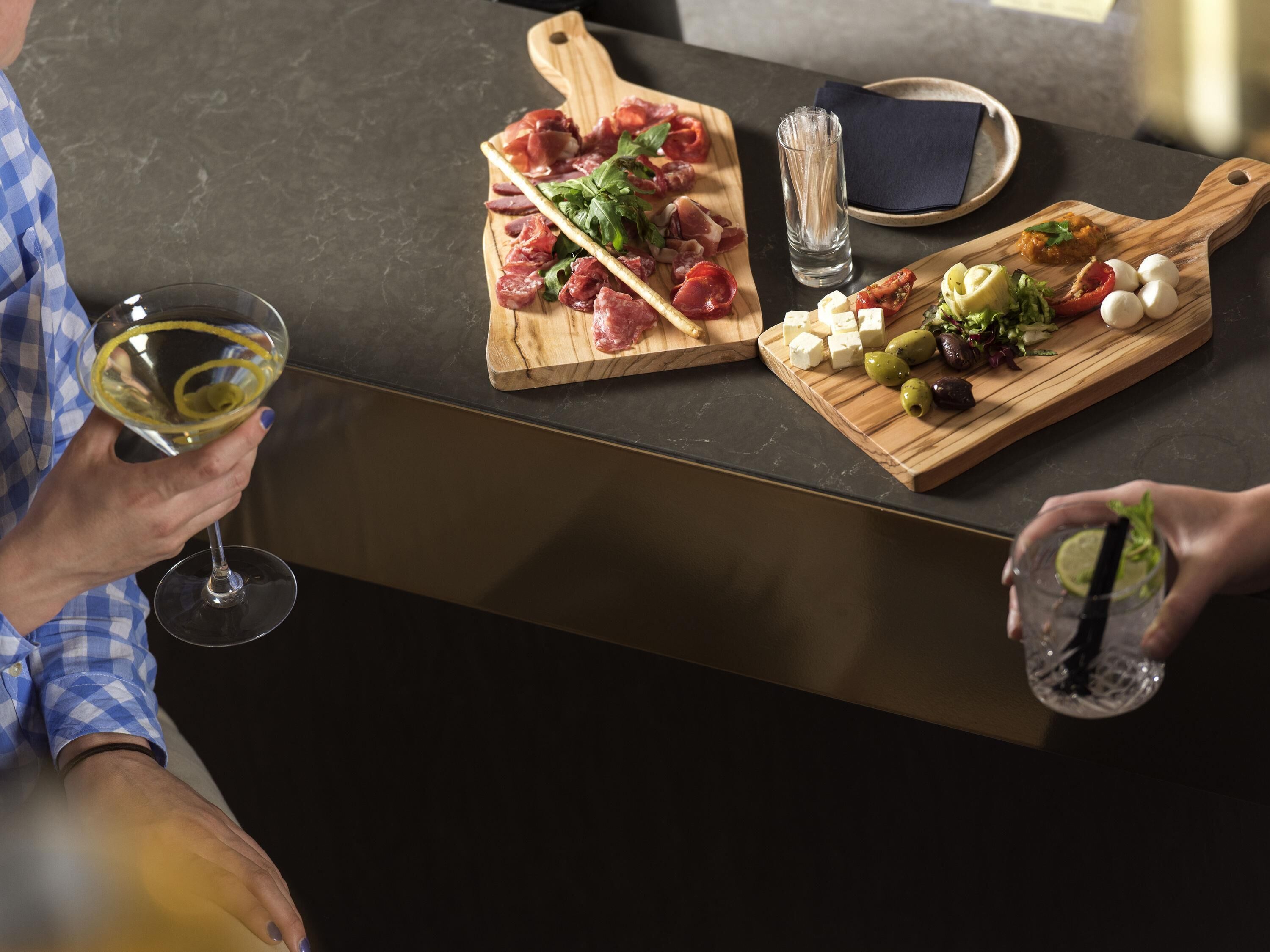 A person holding a wine glass, a wooden board with various cheeses and olives, and another person with a drink.