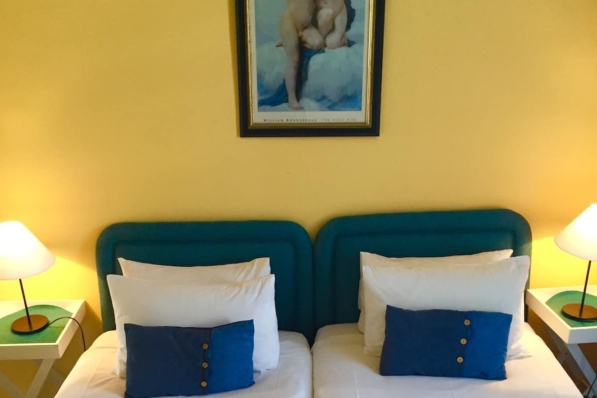 Una cama doble con ropa de cama blanca y azul, dos mesitas de noche con lámparas y un cuadro en la pared.