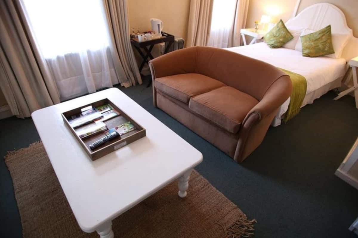 Habitación de hotel con una cama, un sofá marrón, una mesa blanca con una bandeja de libros y una mesita de noche con una lámpara.