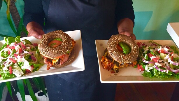 Una persona sostiene dos platos de comida, uno con una hamburguesa y otro con una ensalada, en un restaurante.