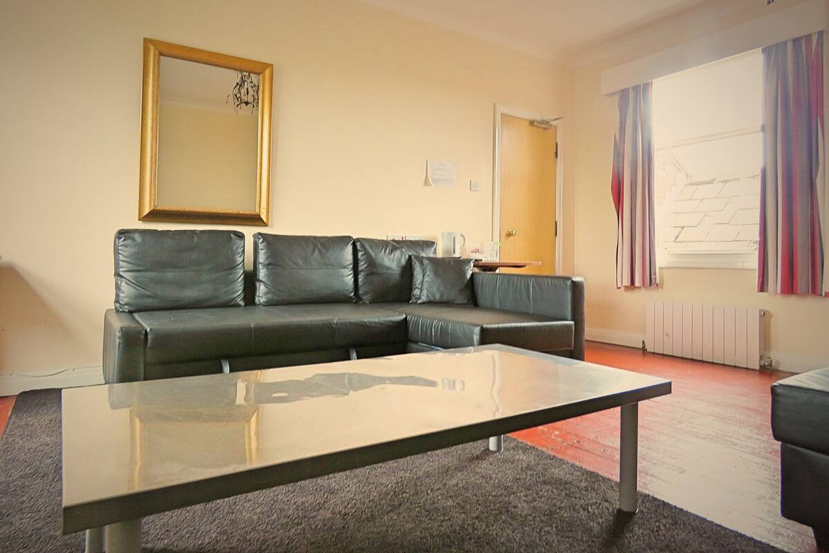 A living room with a black leather sofa, a glass coffee table, a framed picture on the wall, a door, and a window with curtains.