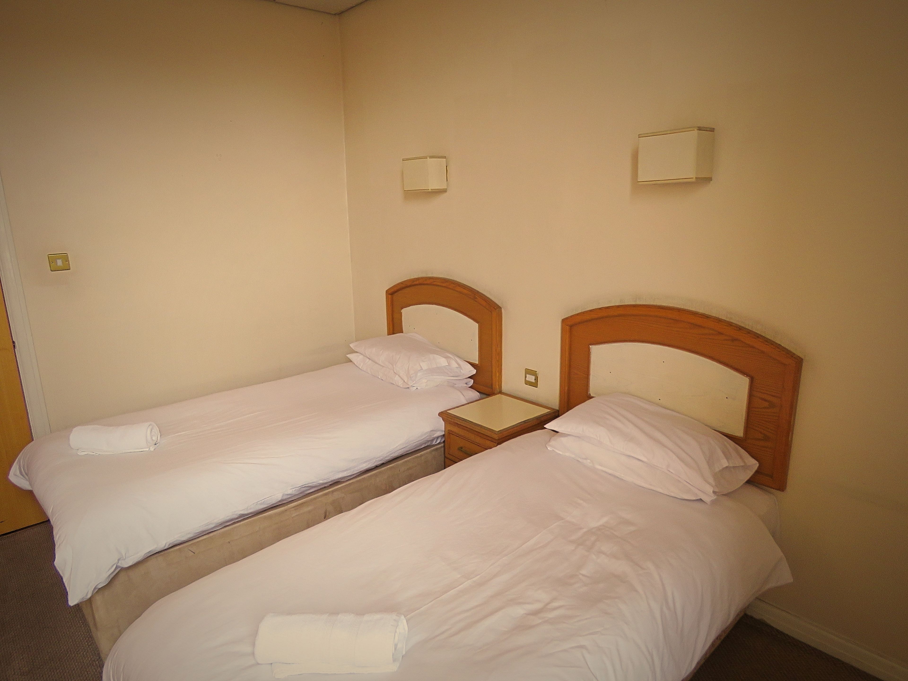Two single beds with wooden headboards and white linens, a bedside table, and a wall-mounted light fixture.