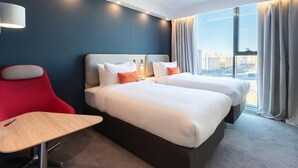 Ein Hotelzimmer mit zwei Betten, einem Schreibtisch, einem Stuhl, einer Lampe und einem großen Fenster mit Vorhängen.