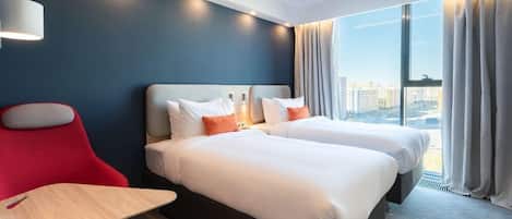 Een hotelkamer met twee bedden, een bureau, een stoel, een lamp en een groot raam met gordijnen.
