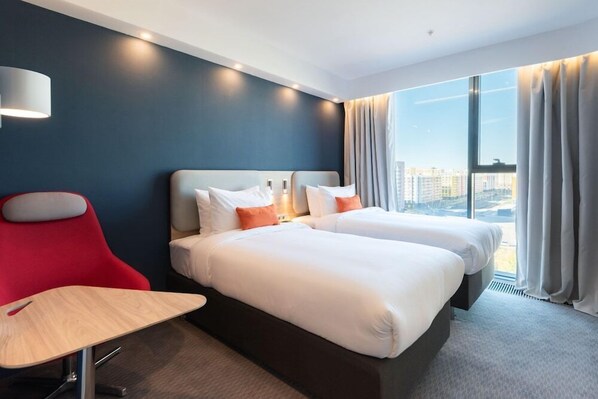 Habitación de hotel con dos camas, un escritorio, una silla, una lámpara y un amplio ventanal con cortinas.