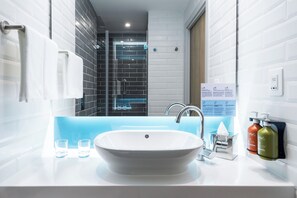 Un baño moderno con un lavamanos blanco, dos vasos, un perchero para toallas, zona de ducha e información en un cartel.