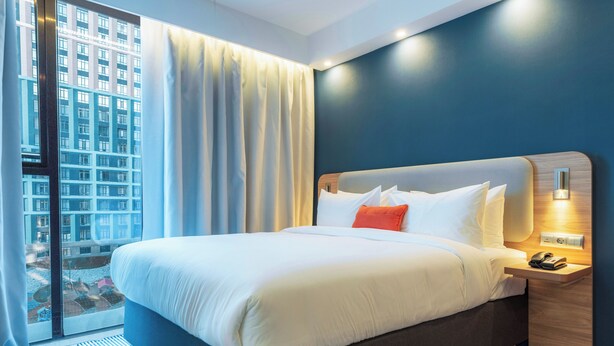 Ein Hotelzimmer mit einem großen Bett, einem Nachttisch, einem Telefon und Blick auf ein Gebäude durch das Fenster.