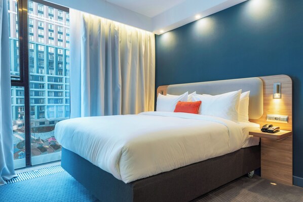 A hotel room with a large bed, a nightstand, a phone, and a view of a building through the window. - Holiday Inn Express Astana - Turan (Astana)