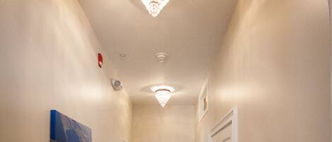 A hallway with wooden flooring, white walls, and a chandelier. There are two framed pictures on the wall and a white door at the end.