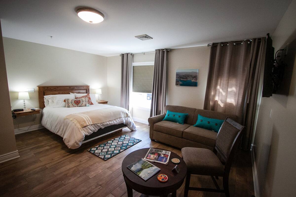 Habitación de hotel con cama, sofá, una mesita con una revista y ventana con cortinas.