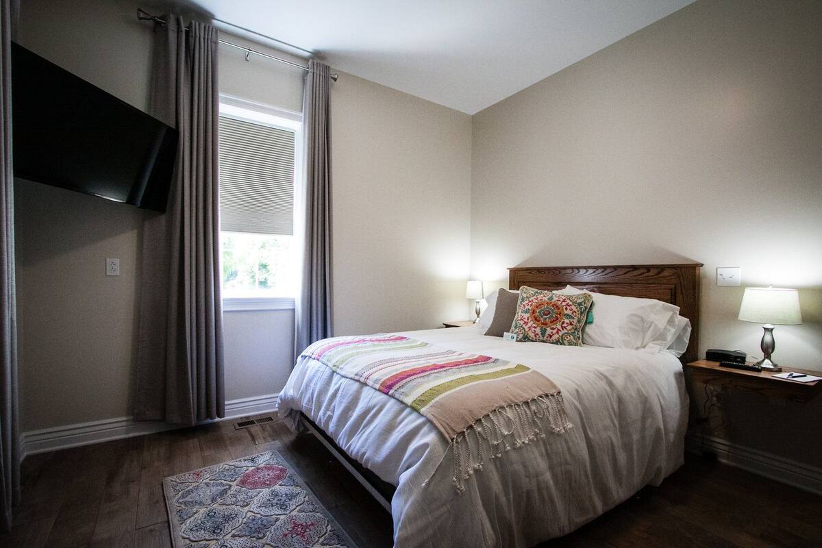 Un dormitorio con una cama, dos lámparas de noche, un televisor montado en la pared y una ventana con cortinas.