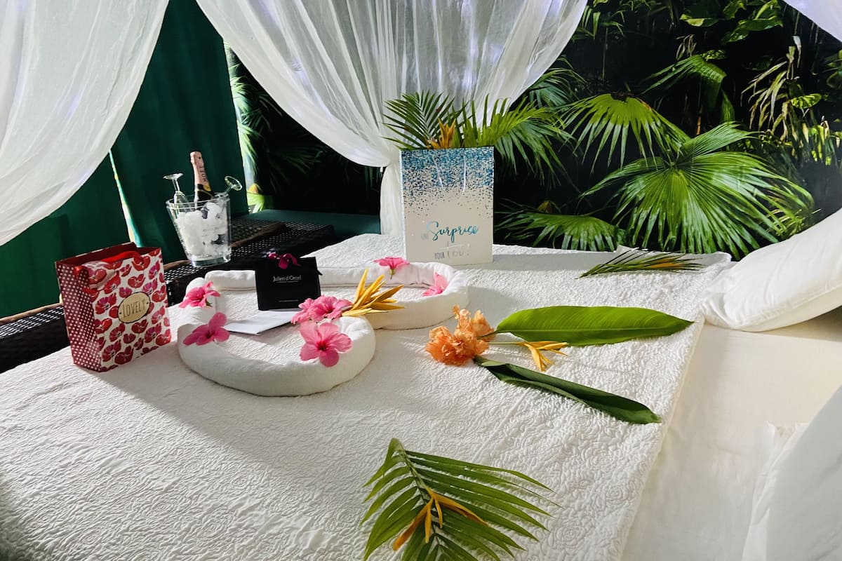 A neatly arranged room with a bed, a small table with a vase of flowers, a gift bag, and a card.