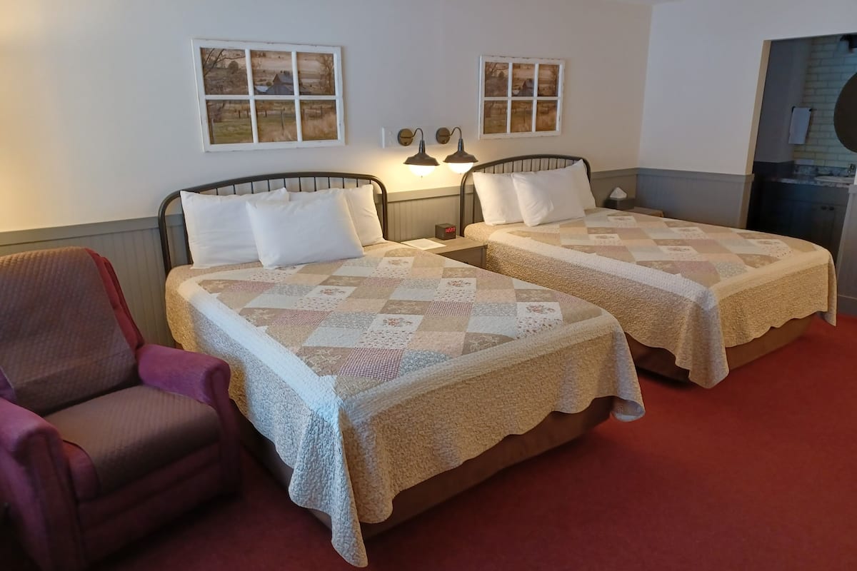 A hotel room with two beds, a chair, and a window with a view of a rural landscape.