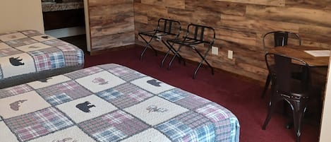 A hotel room with a wooden wall, a TV mounted above a shelf, two black chairs, a desk, and a bed with a plaid quilt.