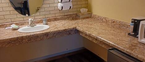 A bathroom with a granite countertop, a sink, a mirror, a towel rack, a tissue box, and a stainless steel refrigerator.