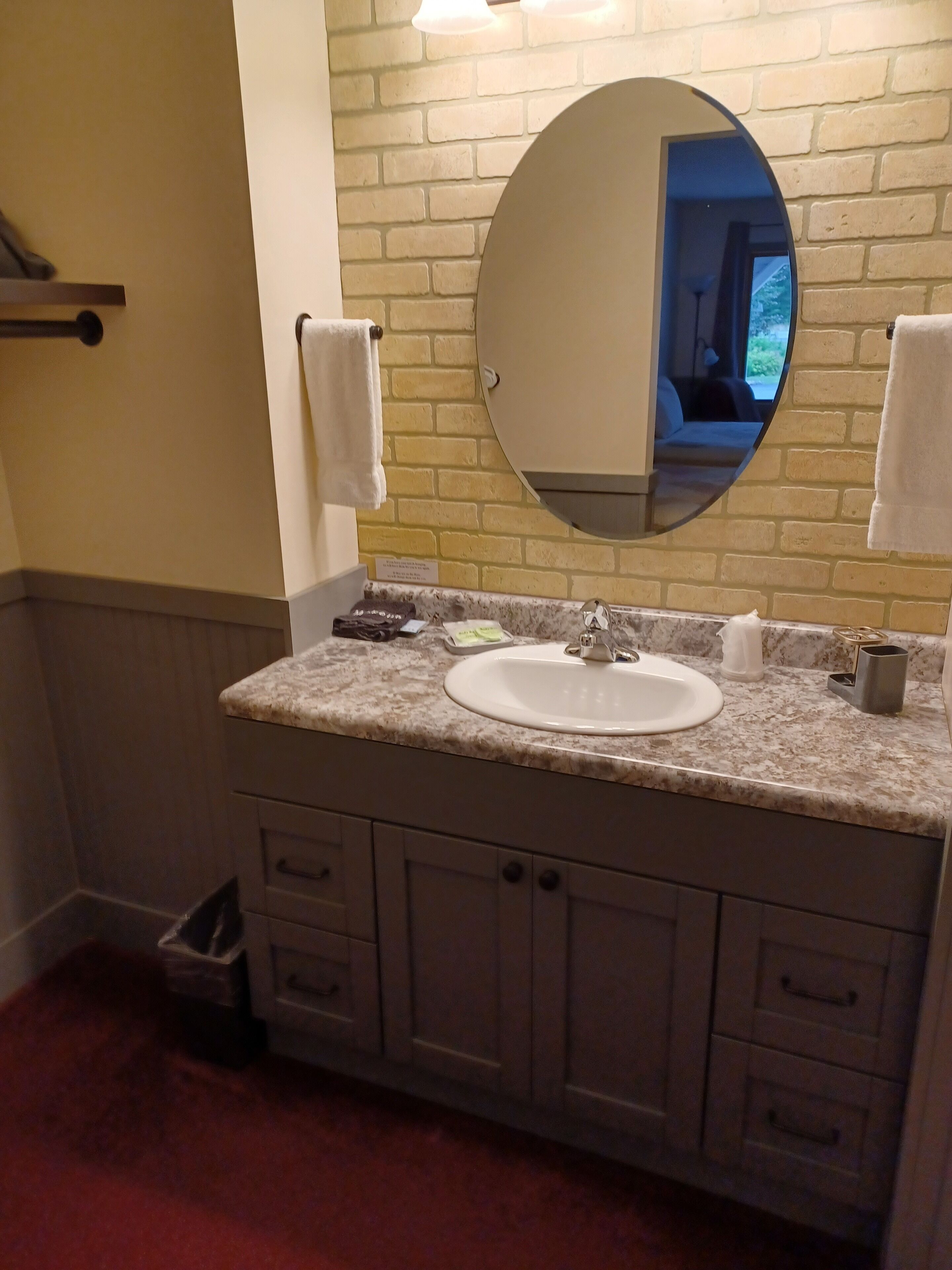 A bathroom with a granite countertop, a white sink, and a round mirror. There are two towels hanging on hooks, a trash bin, and a sign on the wall.