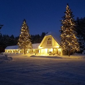 Un paysage enneigé avec une maison illuminée par des lumières chaleureuses, entourée d’arbres couverts de neige.