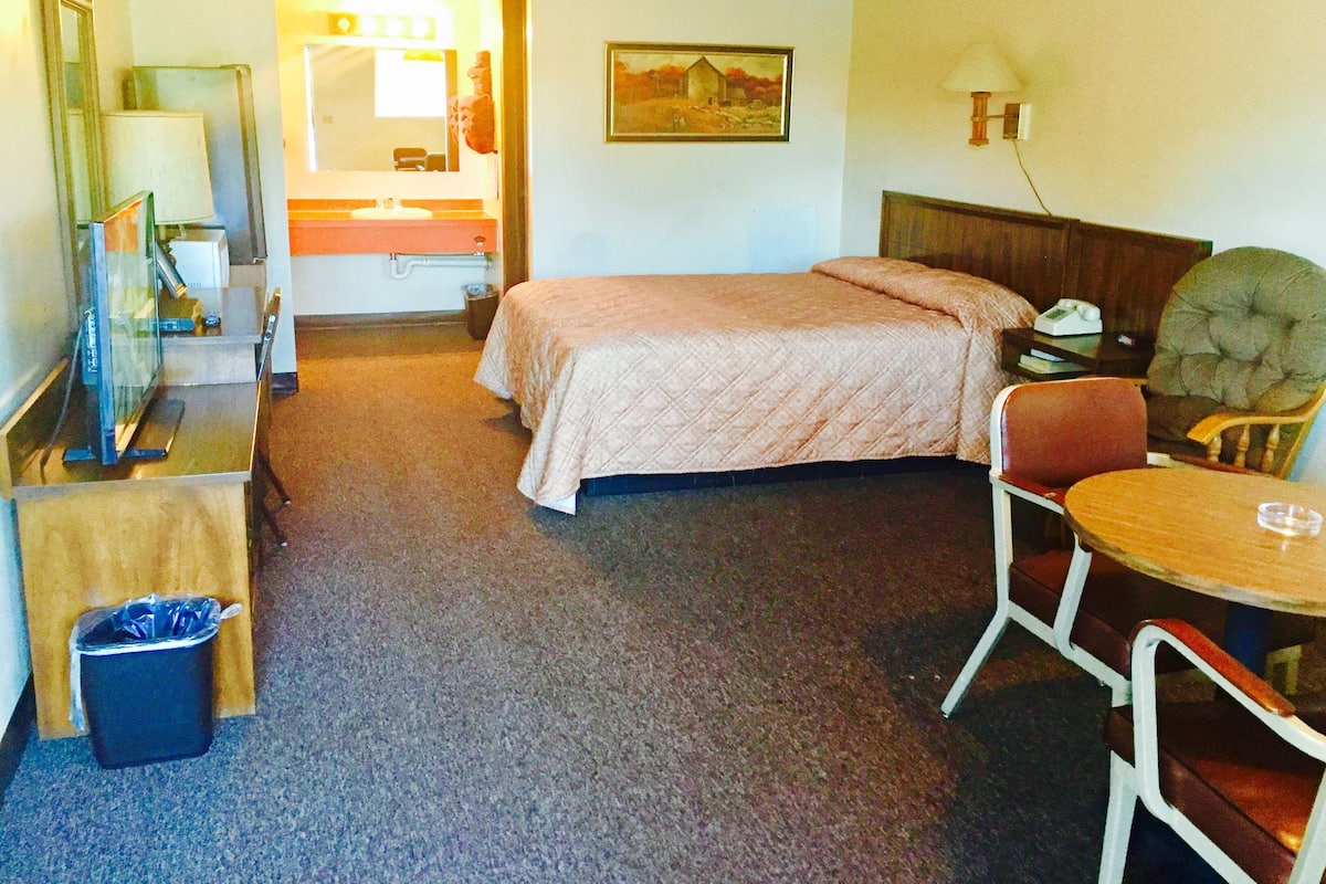 Ett hotellrum med en säng, ett skrivbord med en dator, en stol, ett litet bord, en papperskorg och en tavla på väggen.