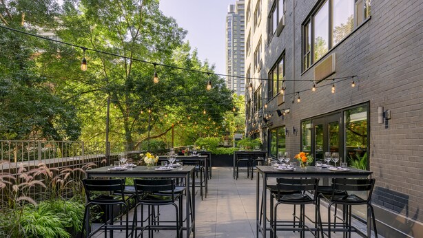 An outdoor dining area with tables and chairs, surrounded by greenery and a building with large windows. - The Anndore House, part of JdV by Hyatt (Toronto)