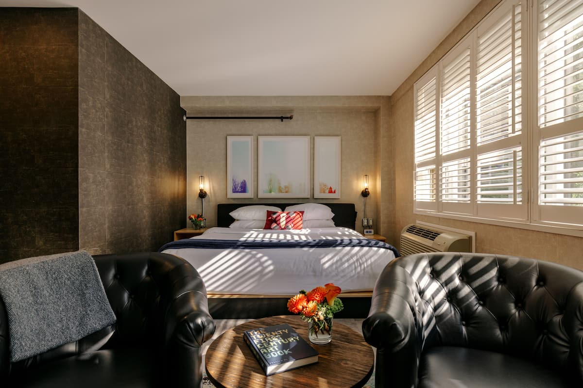 Une chambre d’hôtel moderne avec un grand lit, deux fauteuils en cuir, une petite table ronde avec un livre et des fleurs, et de grandes fenêtres équipées de stores.