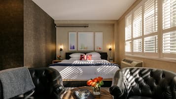 A modern hotel room with a large bed, two leather armchairs, a small round table with a book and flowers, and large windows with blinds.