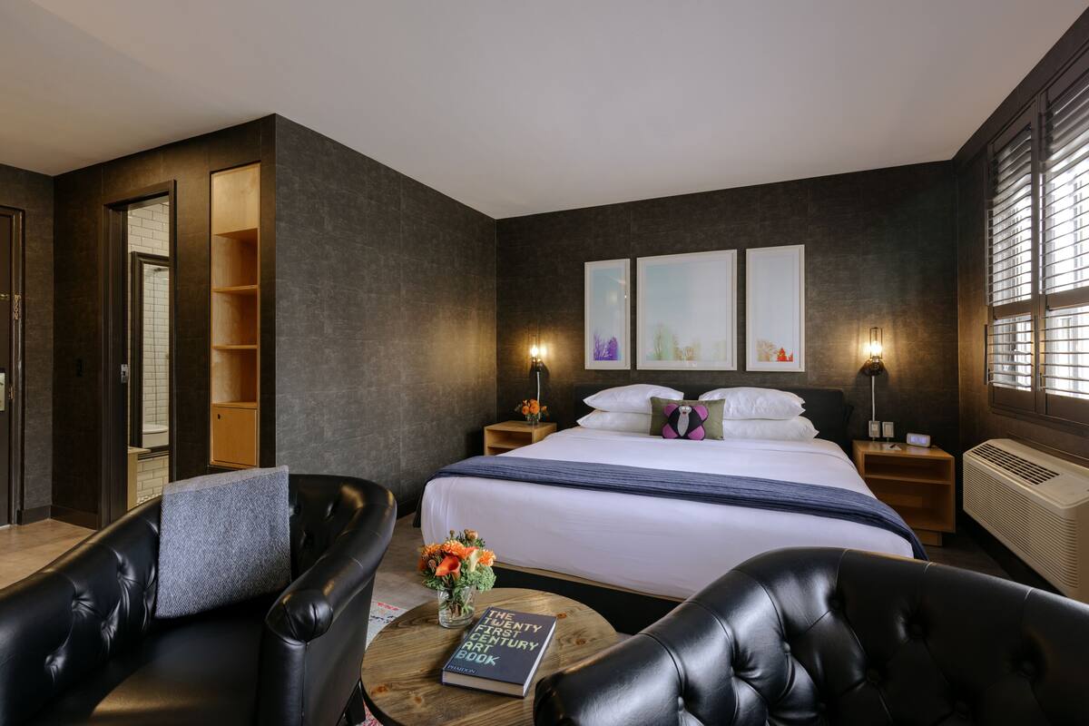 Une chambre d’hôtel moderne avec un grand lit, un canapé en cuir, une petite table ronde avec un livre et des fleurs, et une grande fenêtre avec des stores.