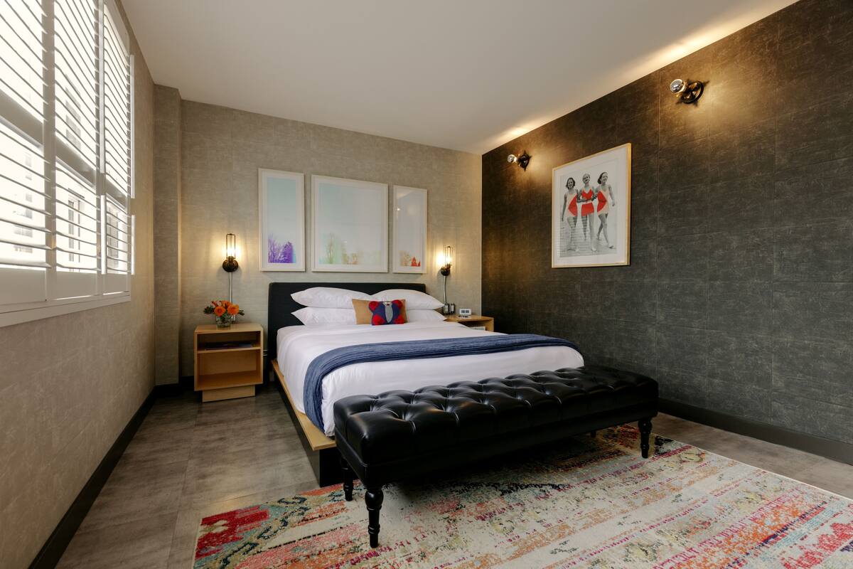 Une chambre à coucher moderne avec un grand lit, un banc noir, un tapis coloré et des œuvres d’art accrochées aux murs.